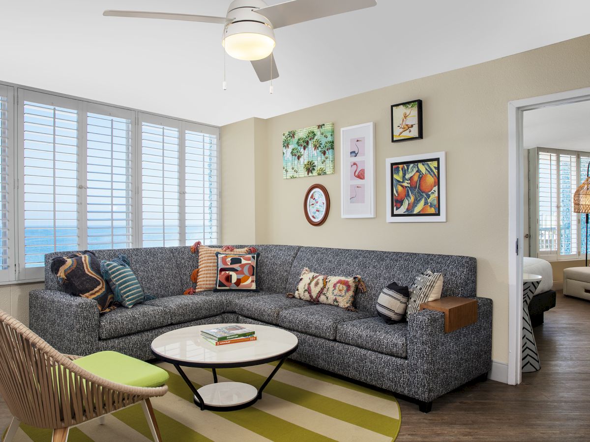 A modern living room with a gray sectional sofa, colorful pillows, a round table, artwork on the wall, and large windows with ocean view.