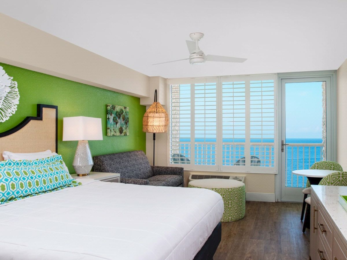 Bright beachside hotel room with a bed, sofa, and ocean view balcony.