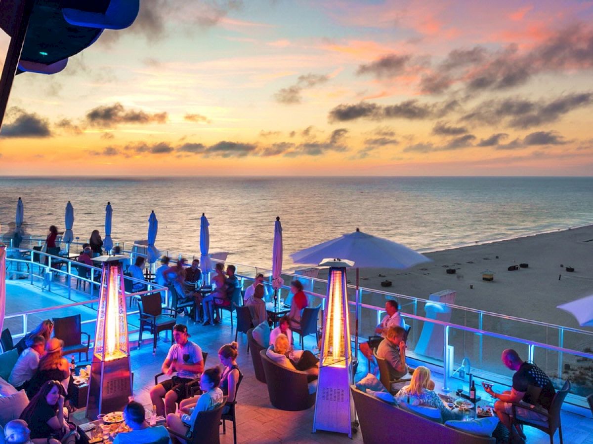 Outdoor terrace with people dining, ocean view, colorful sunset sky, beach in the distance.