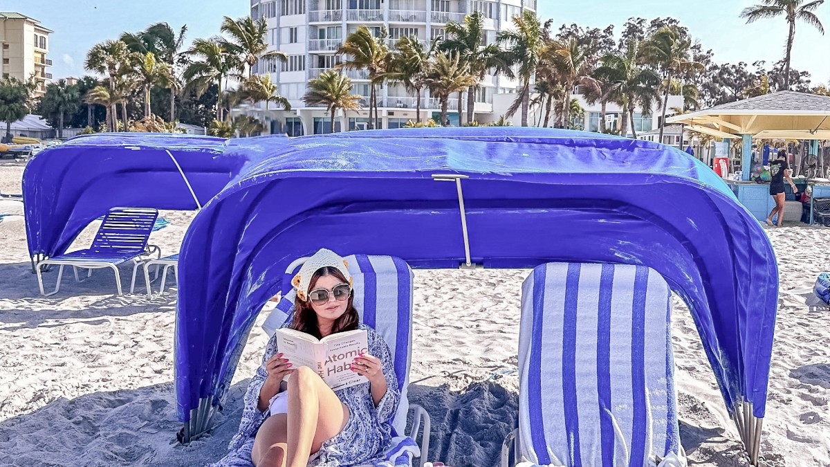 A person is reading on a beach with striped chairs and canopy, buildings in the backdrop.