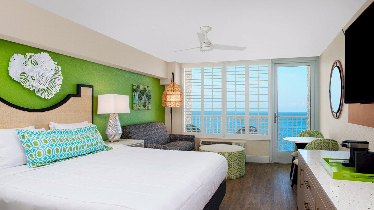 Bright beachside hotel room with a bed, sofa, and ocean view balcony.