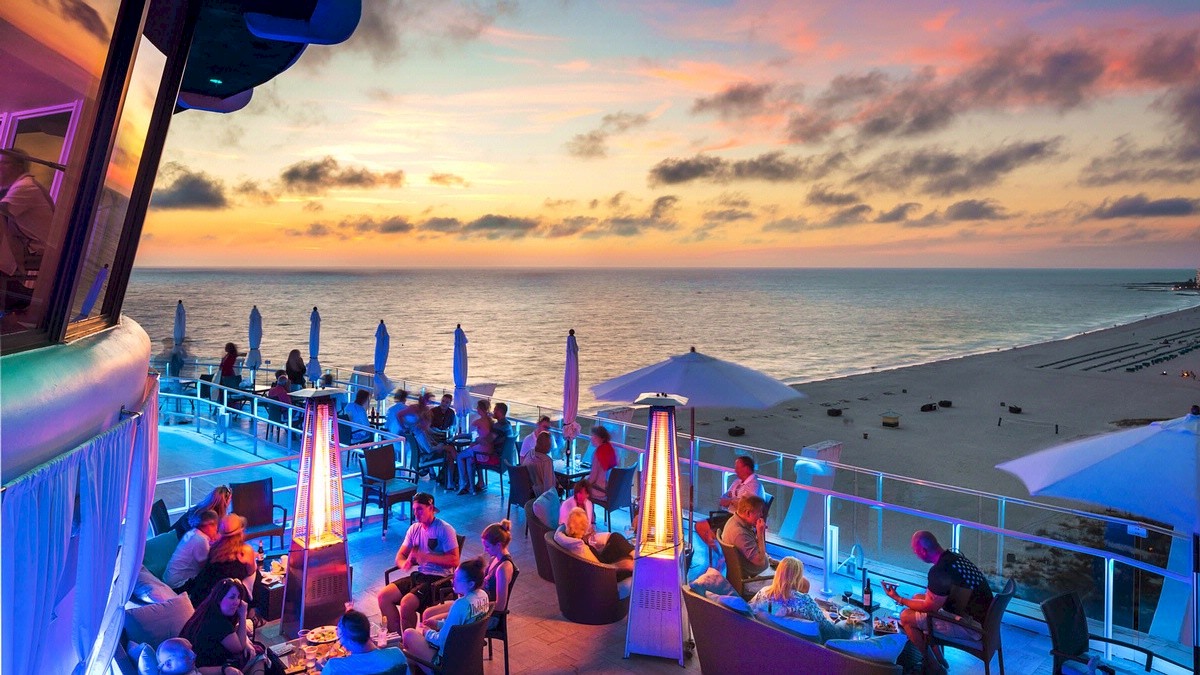 Outdoor terrace with people dining, ocean view, colorful sunset sky, beach in the distance.