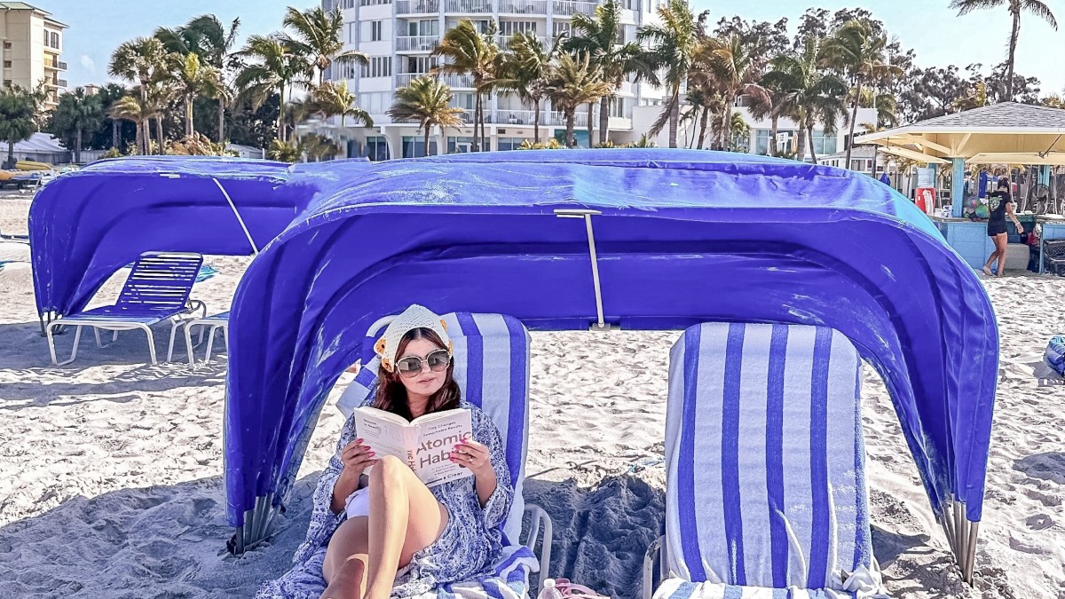 A woman reads on a beach lounge chair under a blue canopy, with a tall, cylindrical building and palm trees in the background.