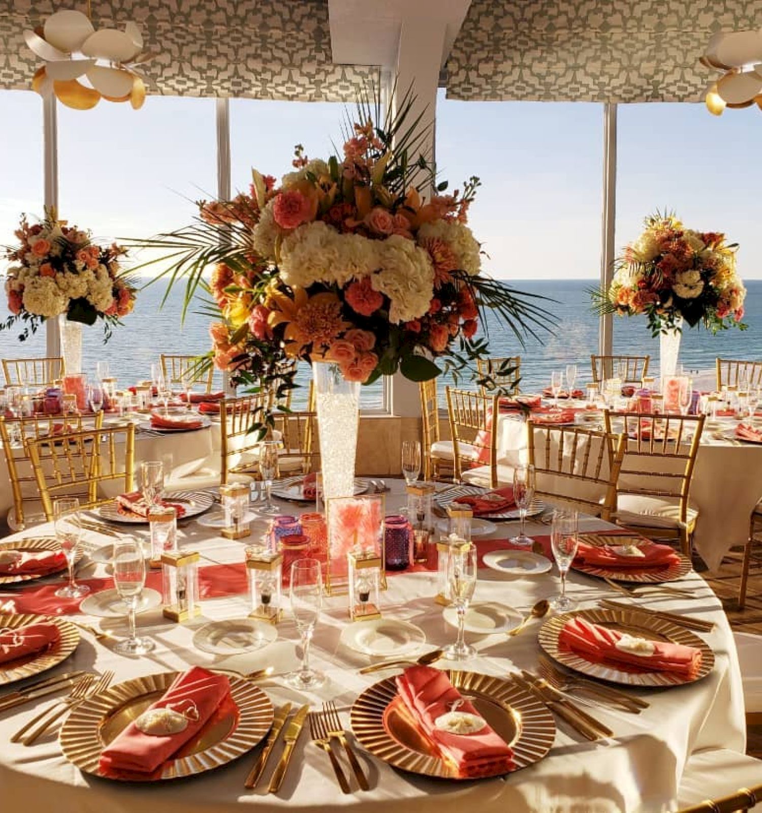 A beautifully decorated banquet hall features round tables with elegant floral centerpieces, gold chairs, and a stunning ocean view through large windows.