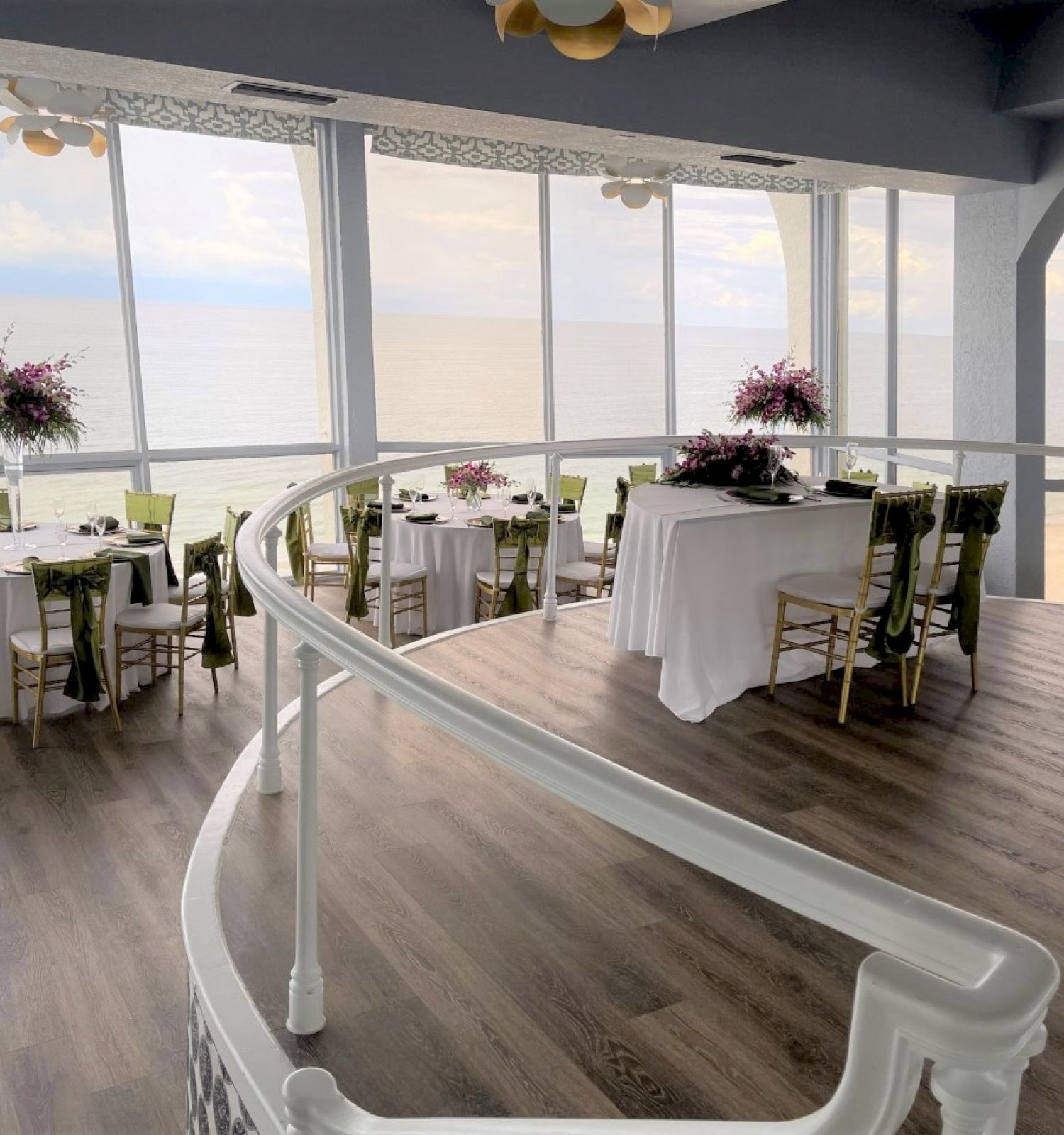 A beautifully decorated dining area overlooking the ocean, featuring tables with flower centerpieces and a curved staircase.
