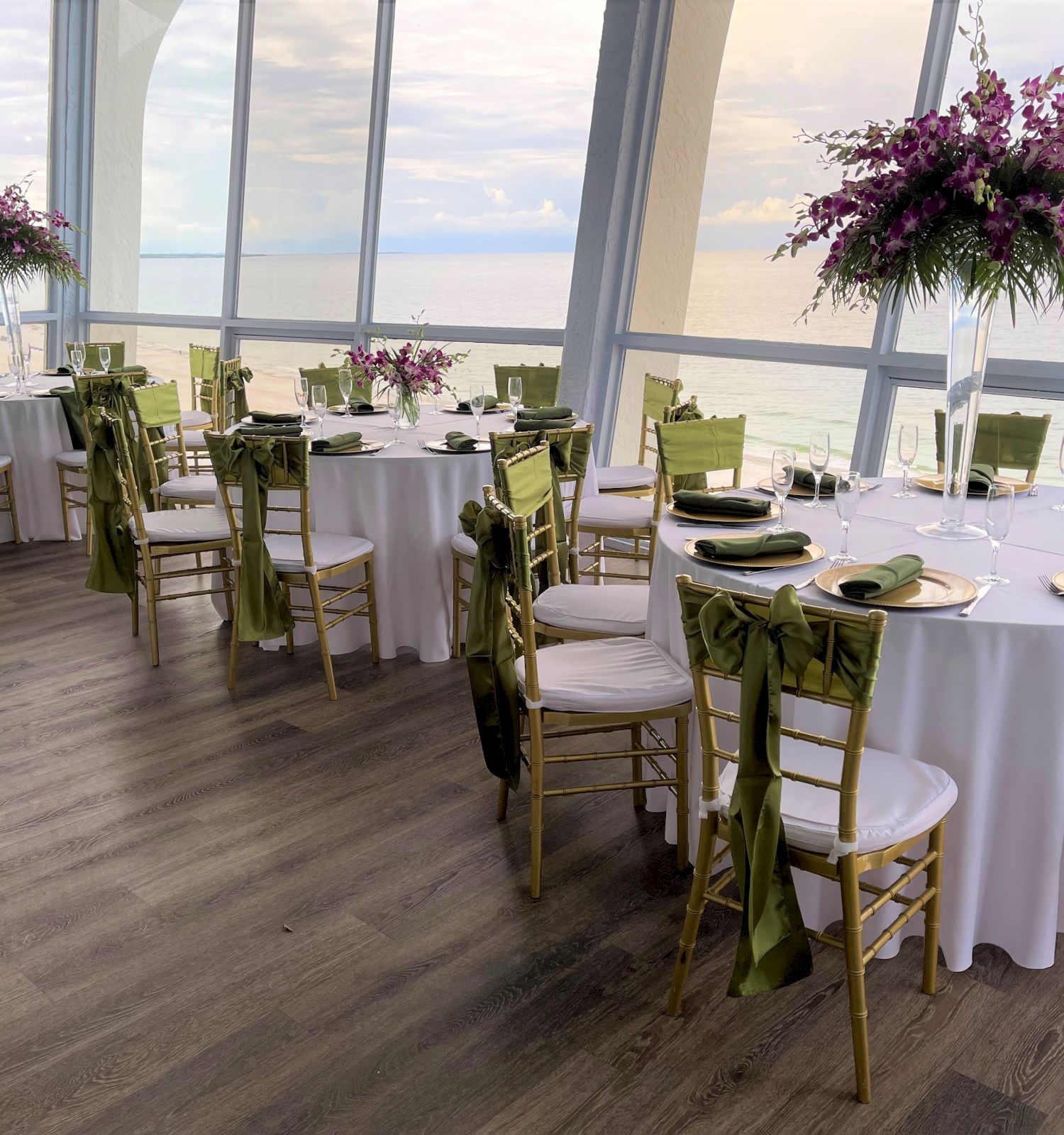 Elegant dining setup in a room with large windows, round tables with white tablecloths, green chairs, and tall floral centerpieces by the sea ending the sentence.