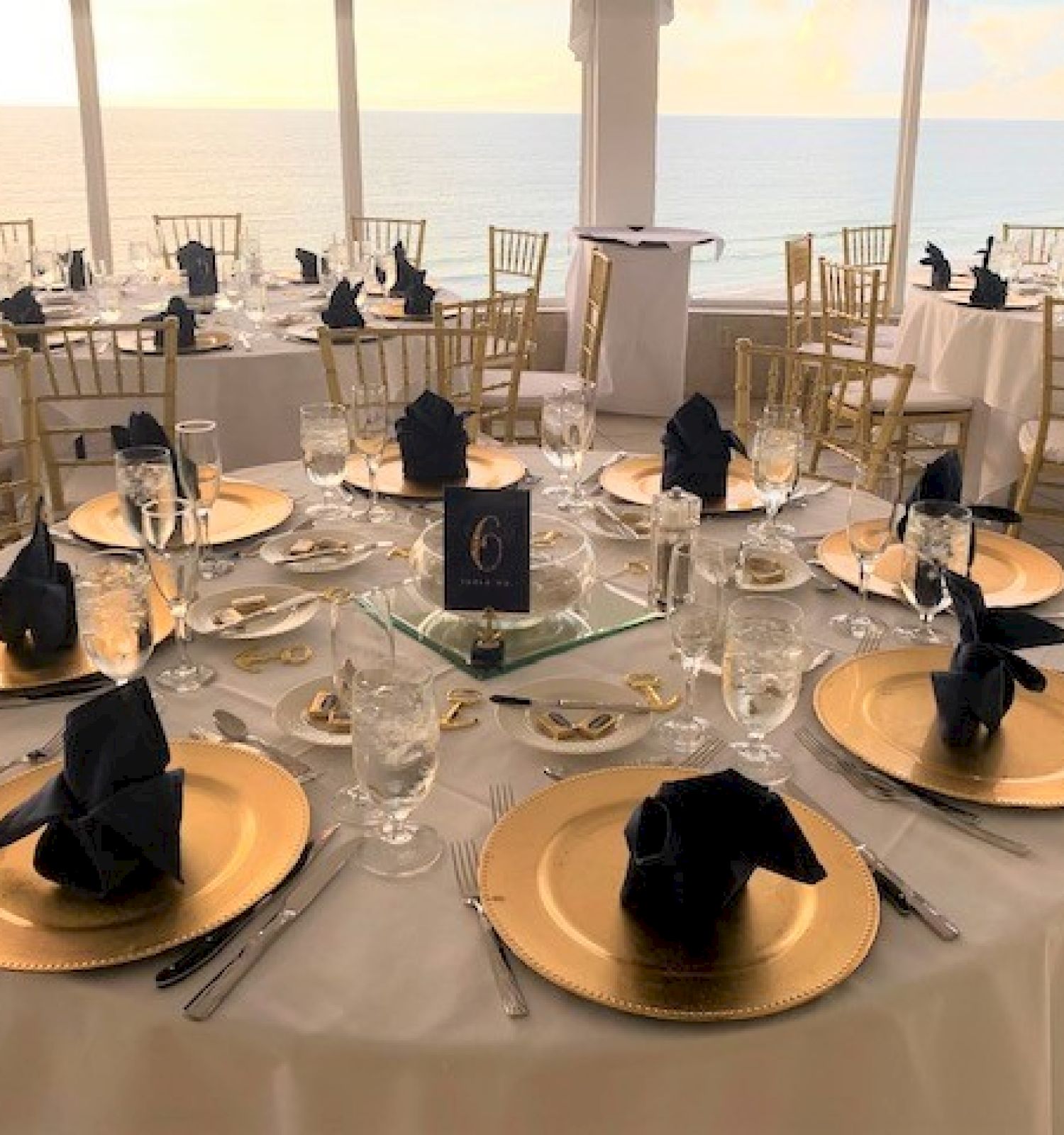 This image shows elegantly set round tables with gold plates, black napkins, and glasses for an event, in a room with a scenic view of the ocean.