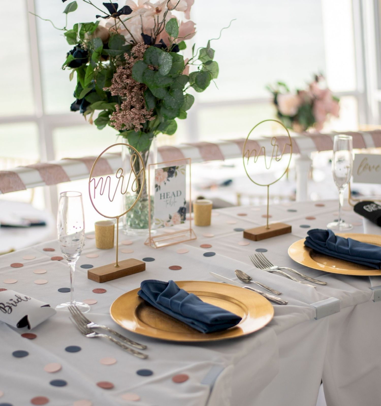 A decorated wedding table features elegant place settings, gold plates, blue napkins, a floral arrangement, and signs reading 