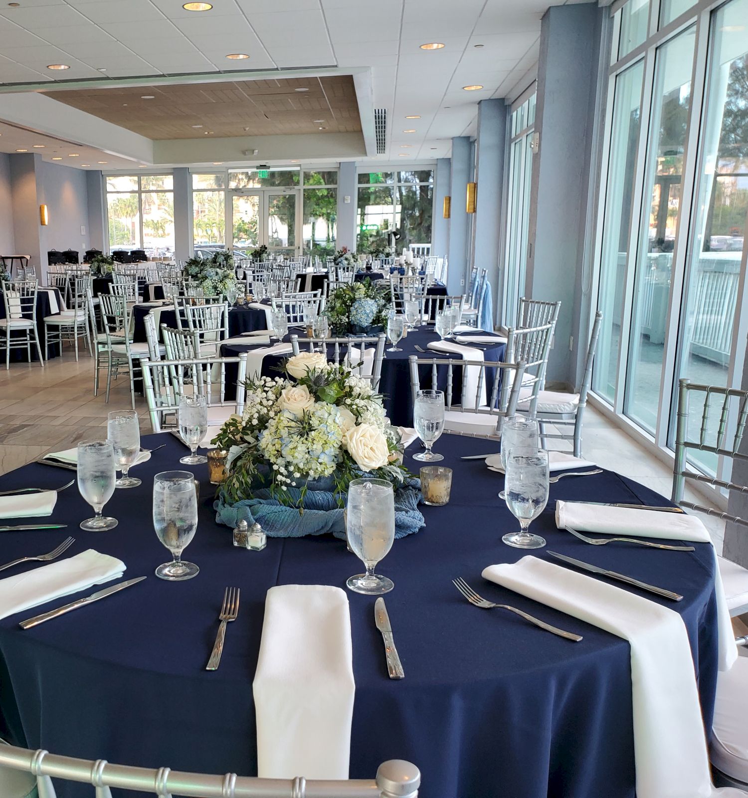 A banquet hall is set up for an event, with round tables covered in navy and white linens, floral centerpieces, and silver chairs around them.