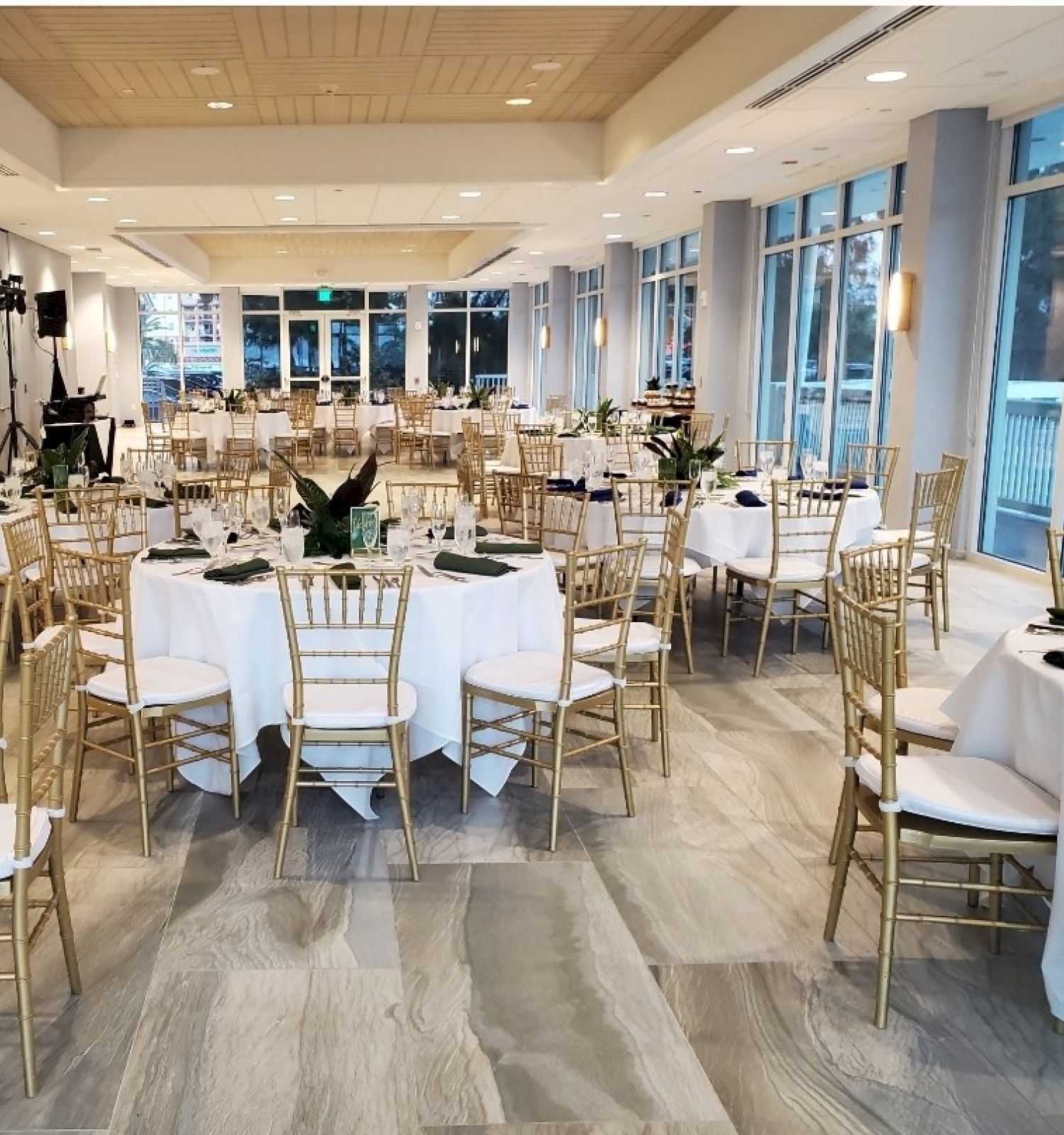 A spacious banquet hall is set up with round tables, white tablecloths, gold chairs, and decorative centerpieces, ready for an event.
