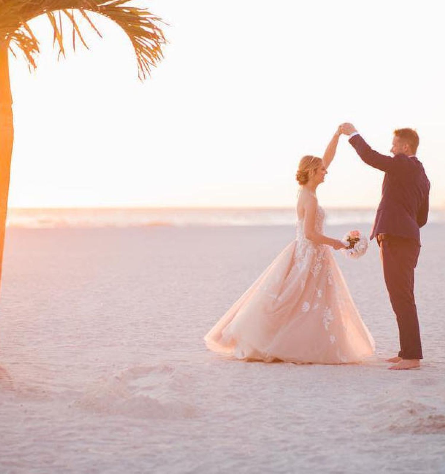 A couple is dancing on a sandy beach at sunset, surrounded by palm trees, with the bride wearing a gown, and the groom in formal attire.