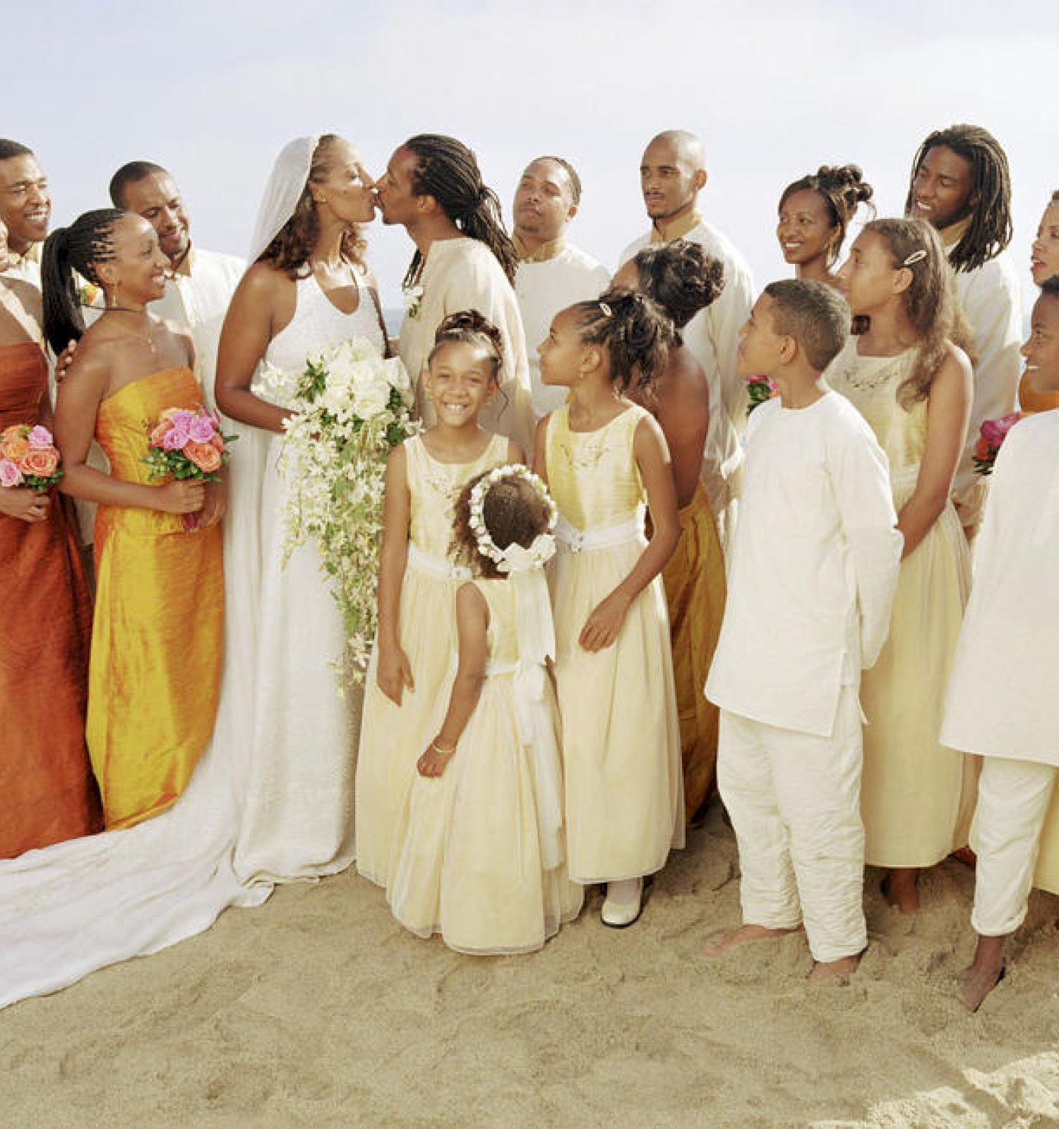 A wedding party posing on a beach with the bride and groom kissing in the center, surrounded by bridesmaids in orange and white dresses, ending the sentence.