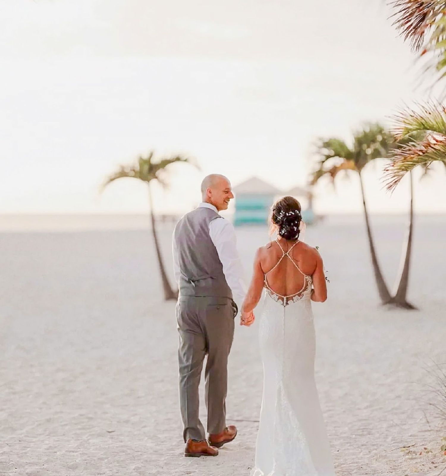 A couple wearing wedding attire walks on a beach adorned with palm trees, heading towards a distant structure, bathed in a serene sunset glow.