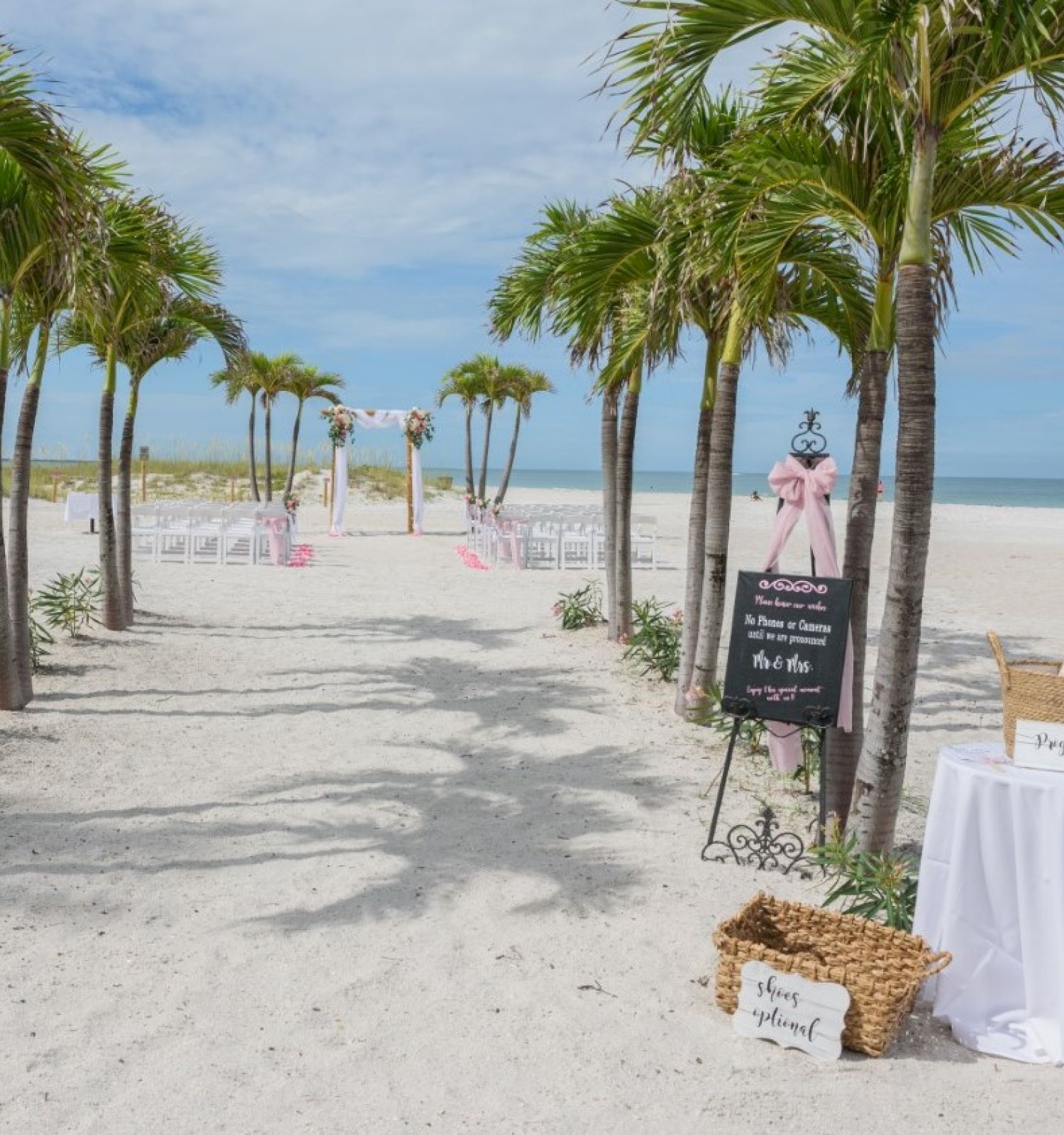 A beach wedding setup with palm trees, a chalkboard sign, and chairs arranged facing the sea. There's a table with a sign-in book and a basket.