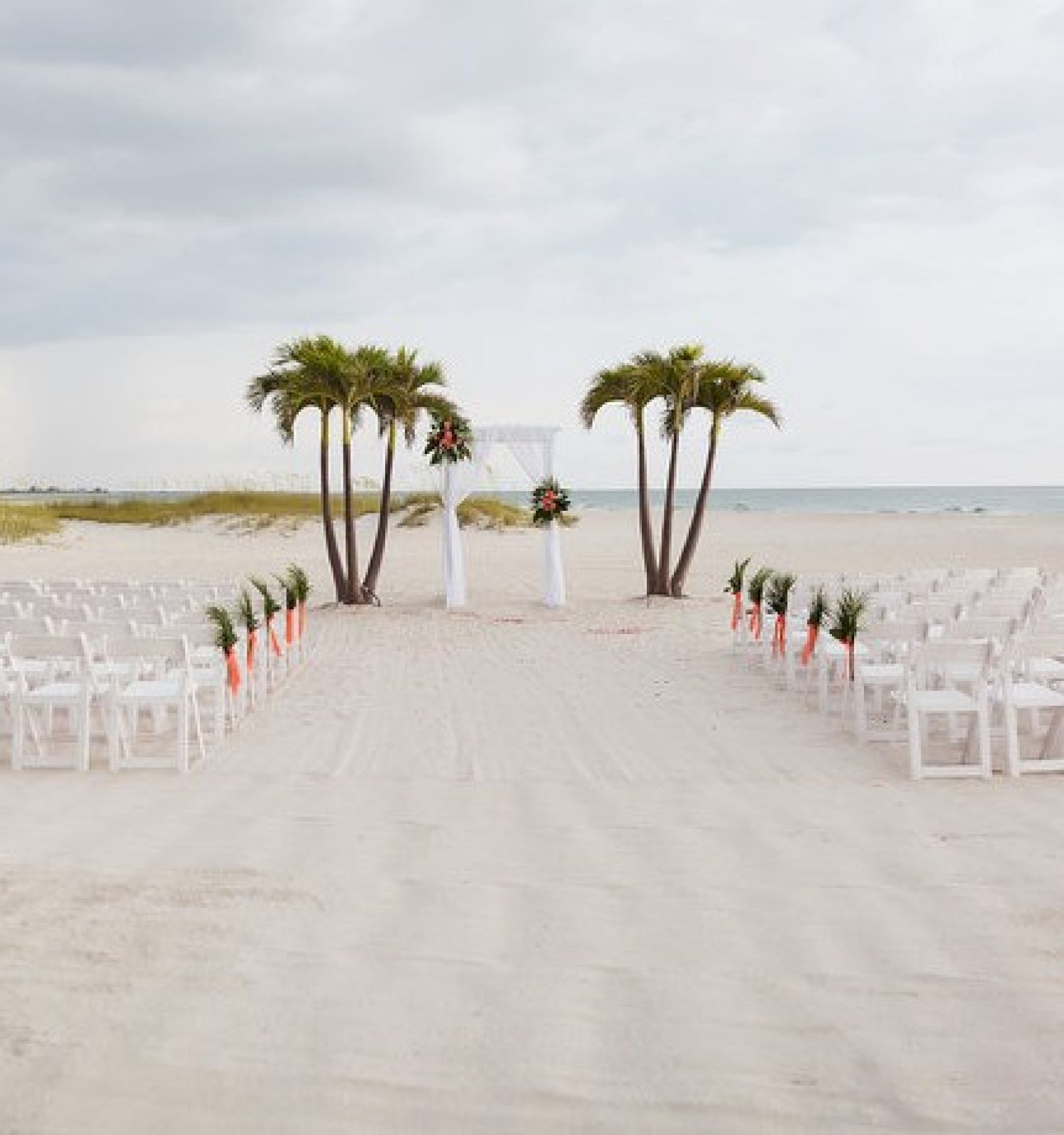 An outdoor wedding setup on a beach with white chairs arranged in rows, facing an arch decorated with flowers and flanked by palm trees, ending the sentence.