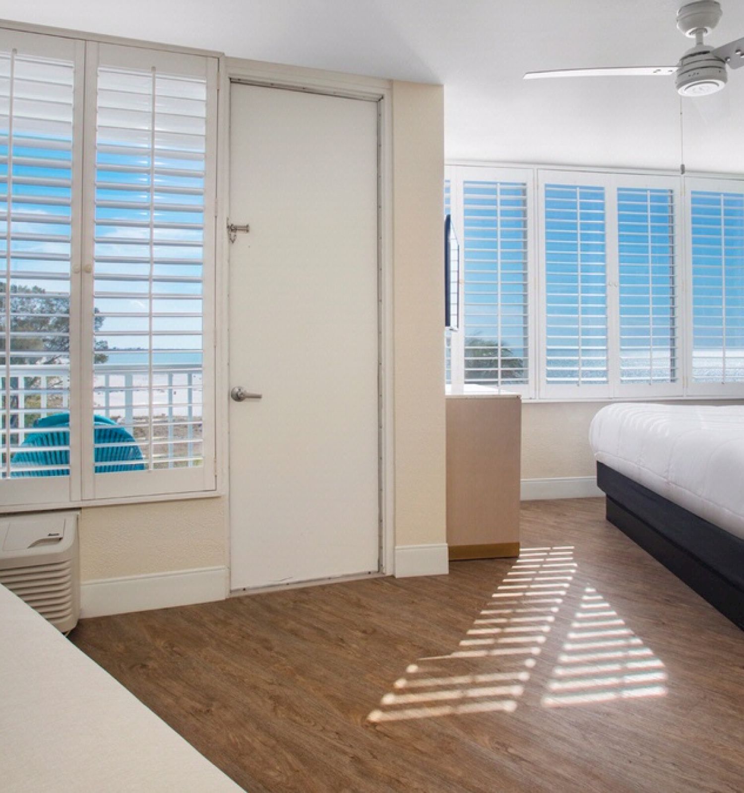 A bright hotel room with wooden flooring, a large bed, colorful pillows, a wicker lamp, a sofa, and large windows with white shutters.