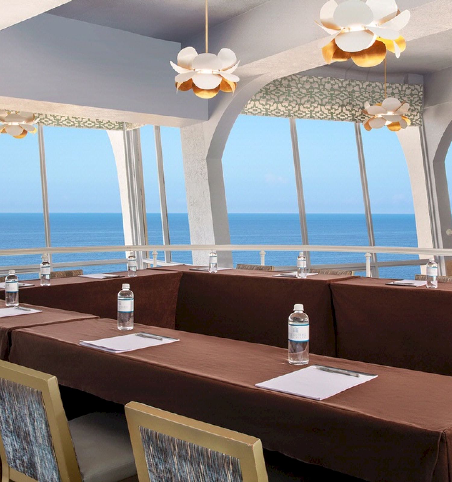 A conference room with ocean views, tables covered in brown cloths, bottled water, notepads, and chairs. Large windows and modern lighting.