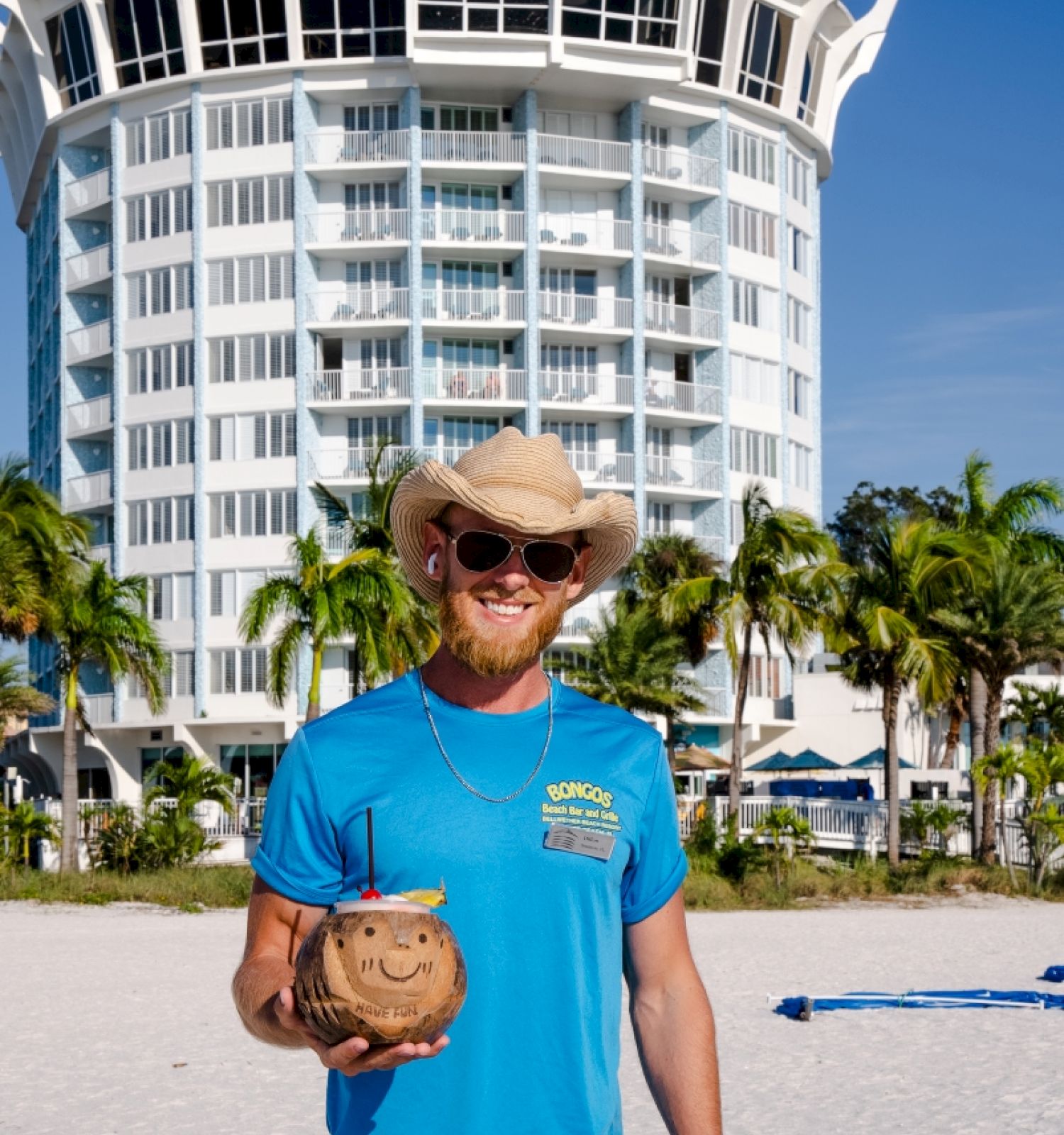 A man in a hat smiles on the beach, holding a coconut, with a hotel in the background.