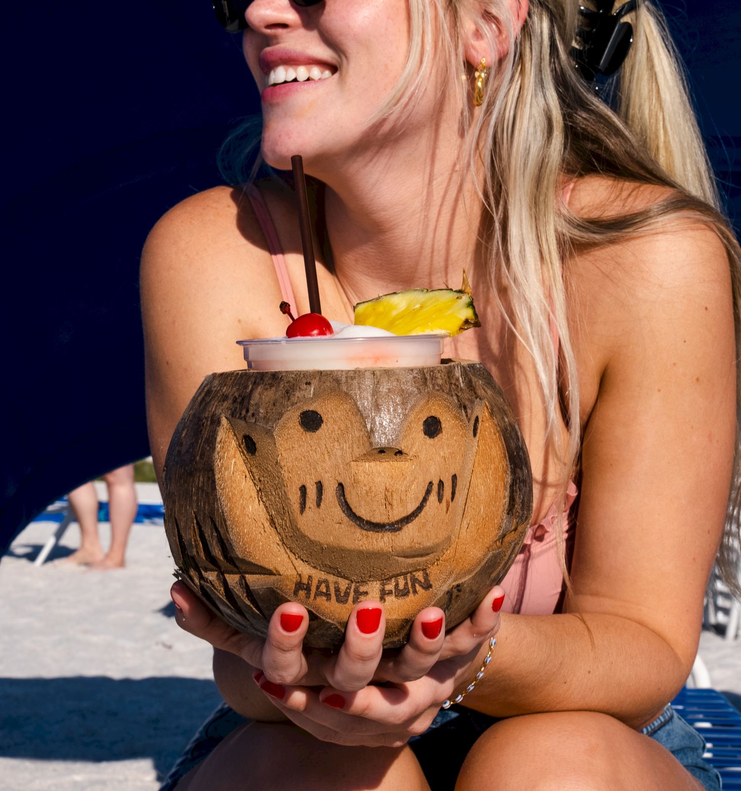 A person is holding a coconut with a smiley face, sitting at a sunny beach.