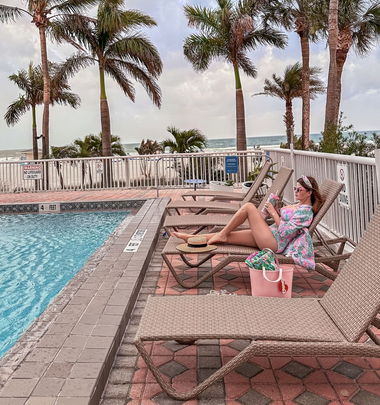Person relaxing by a pool with palm trees and cloudy sky.