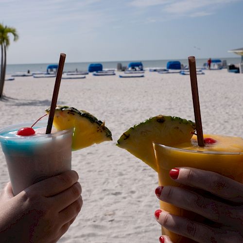 Two hands holding tropical drinks with garnishes on a sandy beach, umbrellas, and a palm tree in the background.