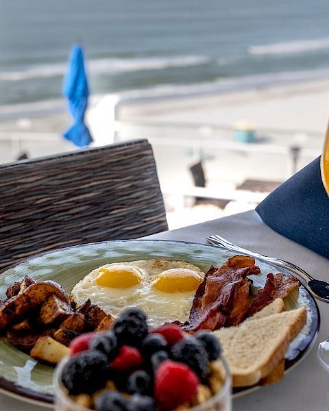 A breakfast plate with eggs, bacon, toast, potatoes, and berries, alongside a glass of iced tea, set on a table overlooking a beach.