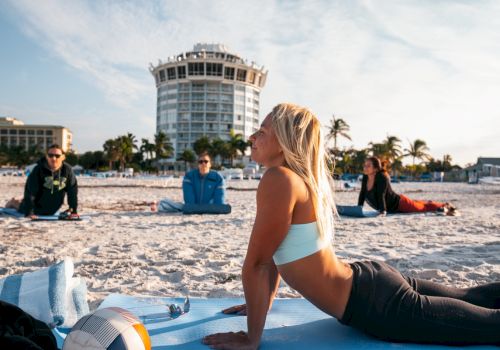 People are doing yoga on a sandy beach with a building and palm trees in the background, and a volleyball in the foreground.