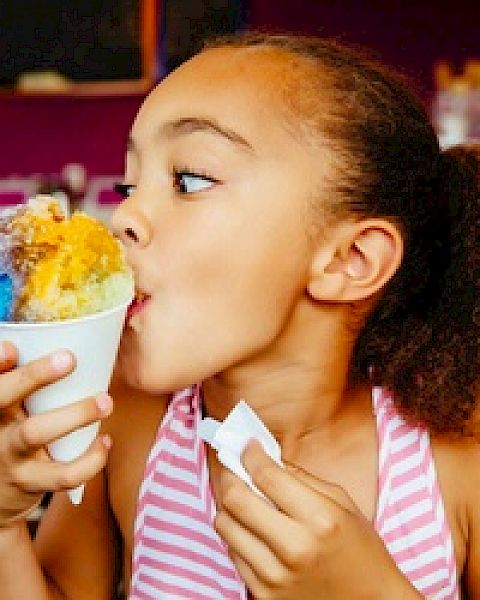 A young girl in a striped shirt enjoys a colorful shaved ice treat, holding a napkin in one hand, against a vibrant background.