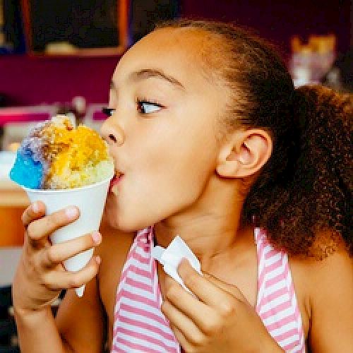 A young girl in a striped shirt enjoys a colorful shaved ice treat, holding a napkin in one hand, against a vibrant background.
