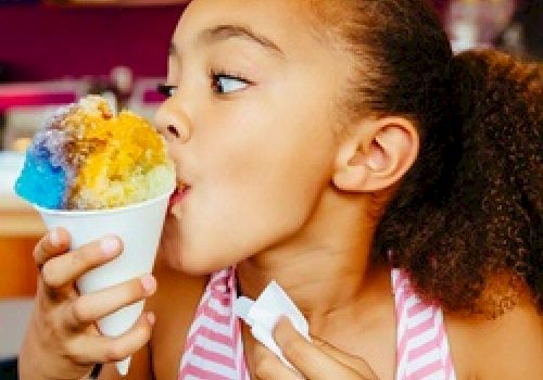 A young girl in a striped shirt enjoys a colorful shaved ice treat, holding a napkin in one hand, against a vibrant background.