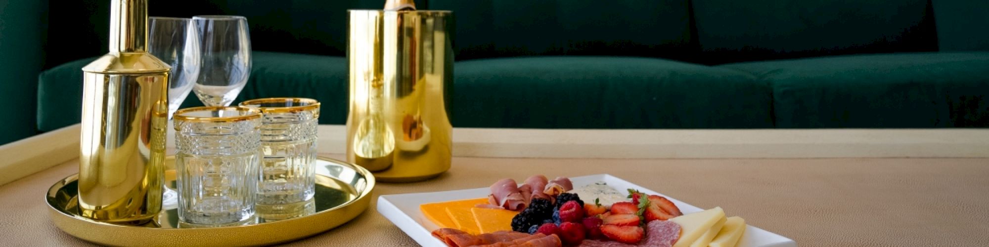 The image shows a tray with gold-colored drinkware and a platter with cheese, meats, and fruits, set in front of a green couch.