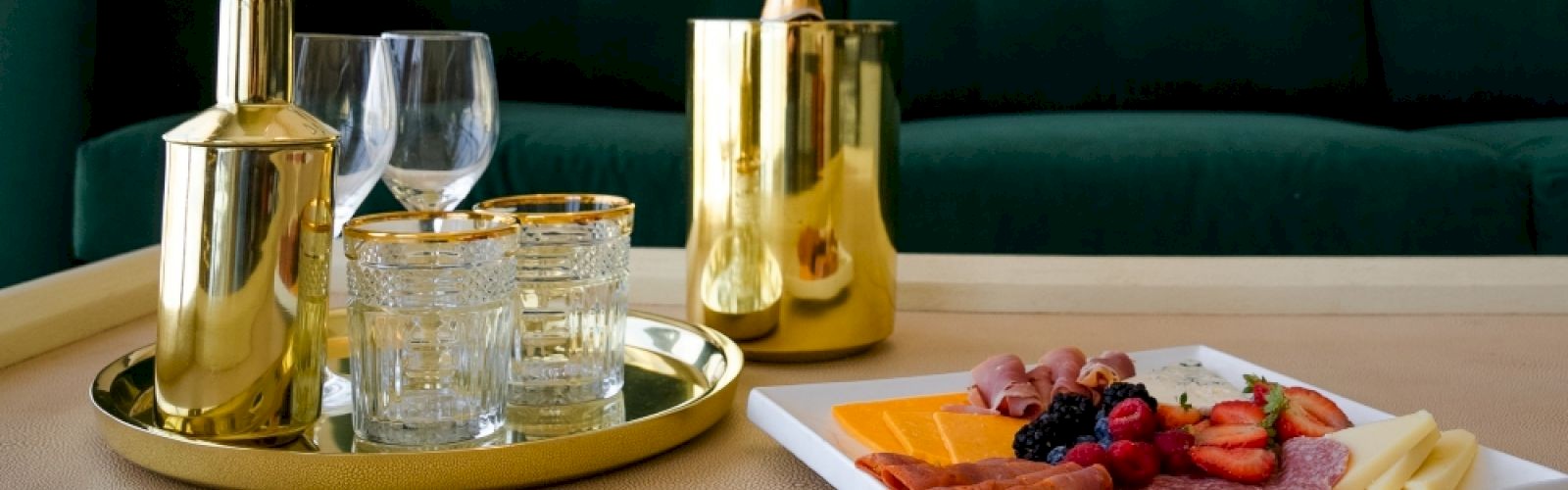 The image shows a tray with gold-colored drinkware and a platter with cheese, meats, and fruits, set in front of a green couch.