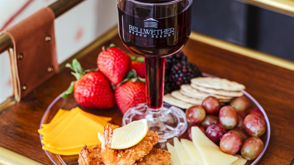 A platter with strawberries, cheese, grapes, crackers, blackberries, and shrimp, accompanied by a glass of red wine on a wooden tray.