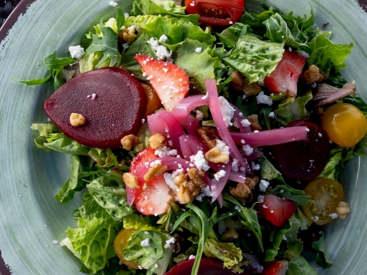 A fresh salad with mixed greens, sliced strawberries, beets, cherry tomatoes, pickled onions, walnuts, and crumbled cheese on a green plate.