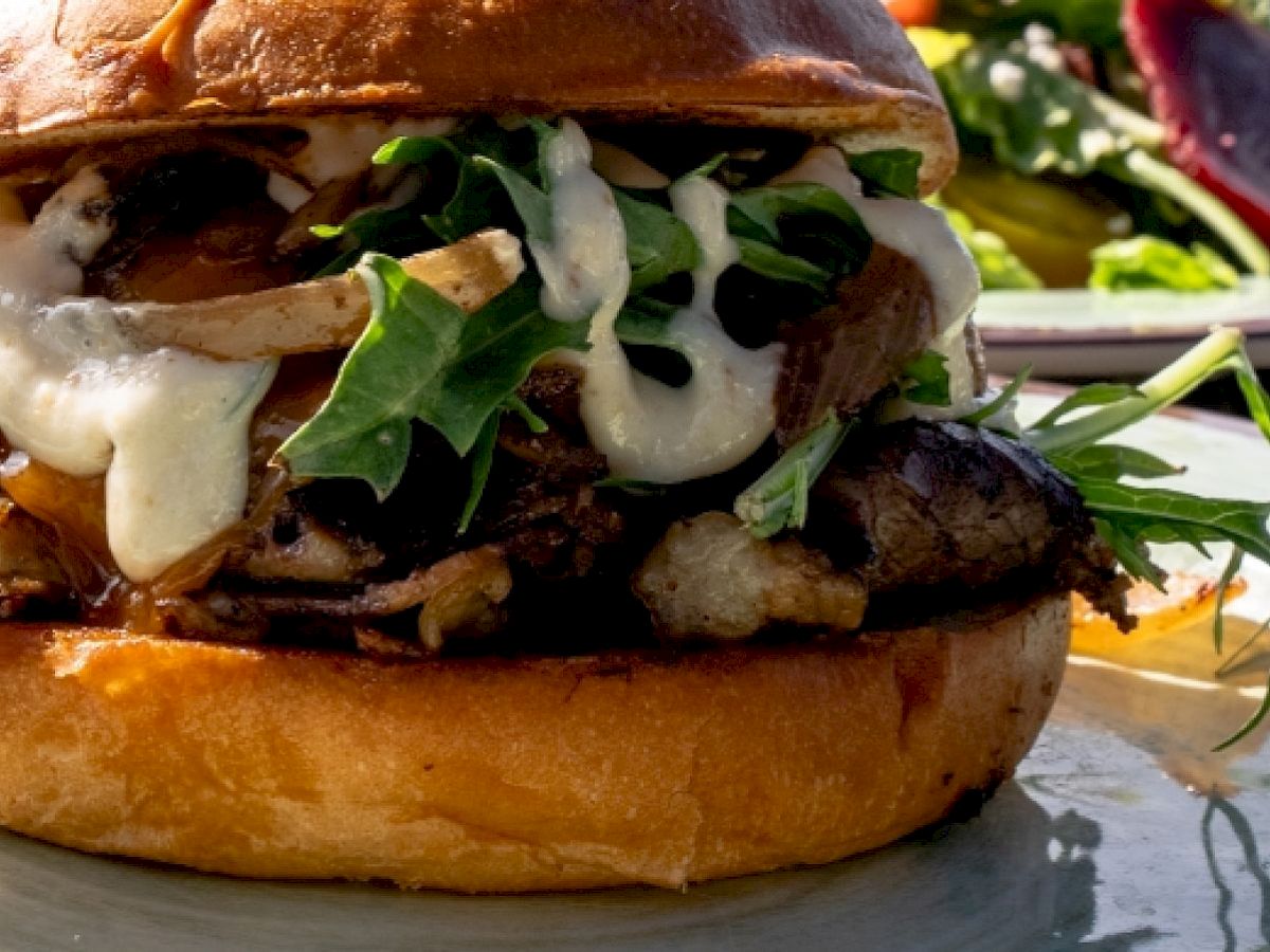 The image shows a close-up of a gourmet burger with meat, leafy greens, sauce, and a toasted bun, with a salad in the background.