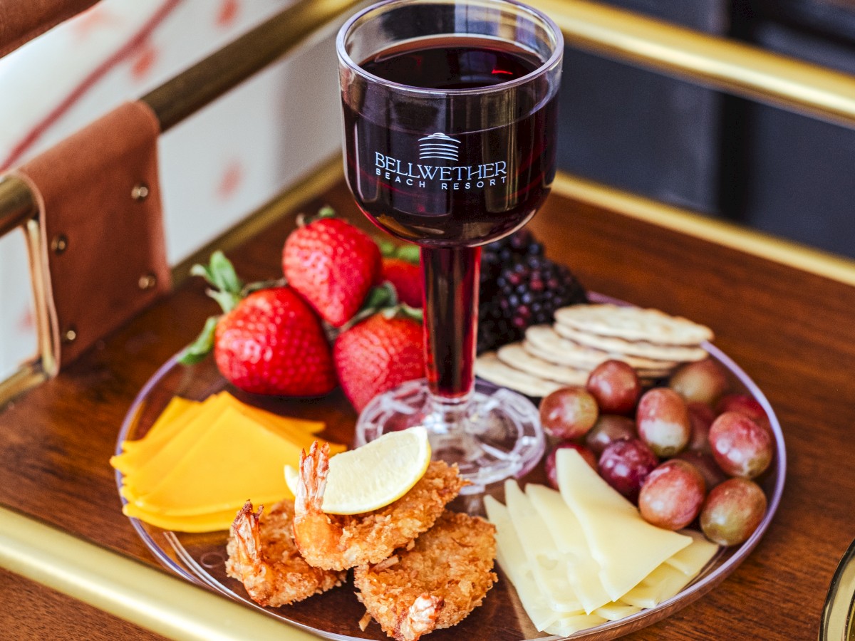 A platter with strawberries, cheese, grapes, crackers, blackberries, and shrimp, accompanied by a glass of red wine on a wooden tray.