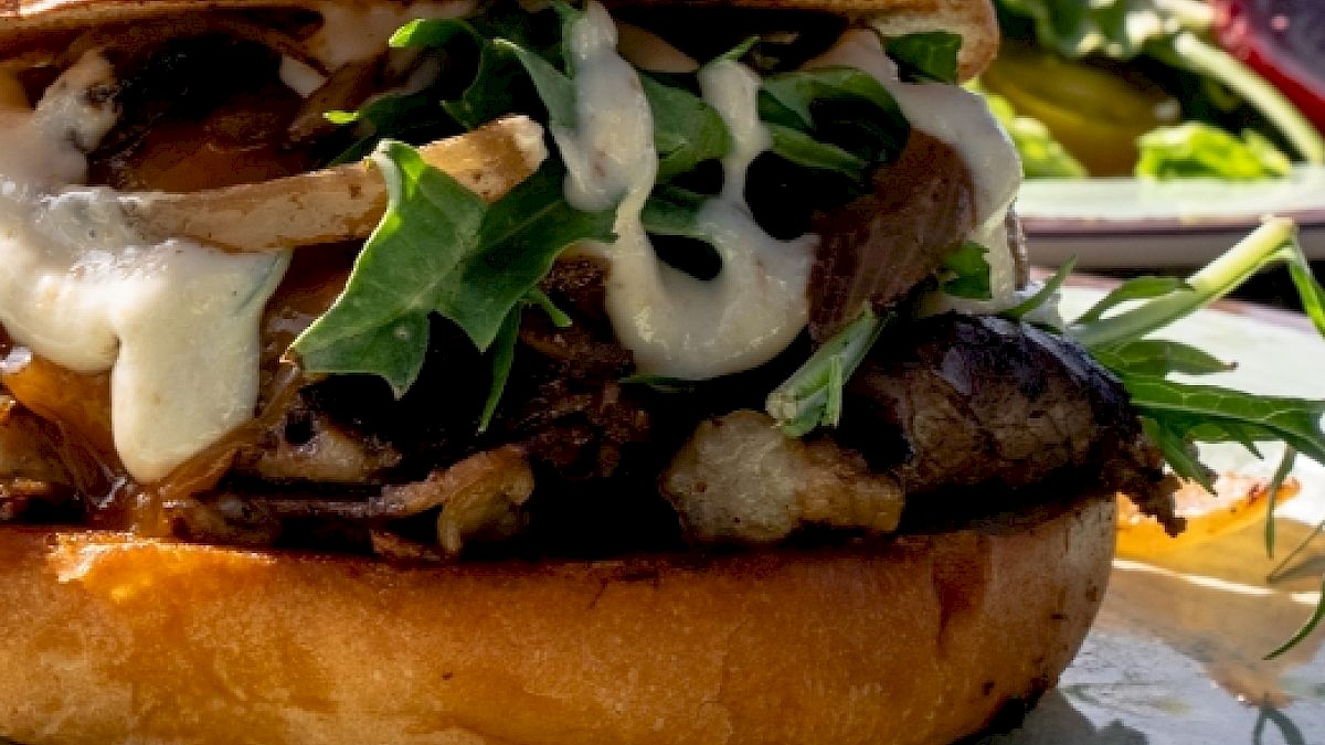 The image shows a close-up of a gourmet burger with meat, leafy greens, sauce, and a toasted bun, with a salad in the background.