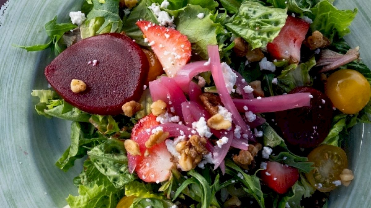 A fresh salad with mixed greens, sliced strawberries, beets, cherry tomatoes, pickled onions, walnuts, and crumbled cheese on a green plate.