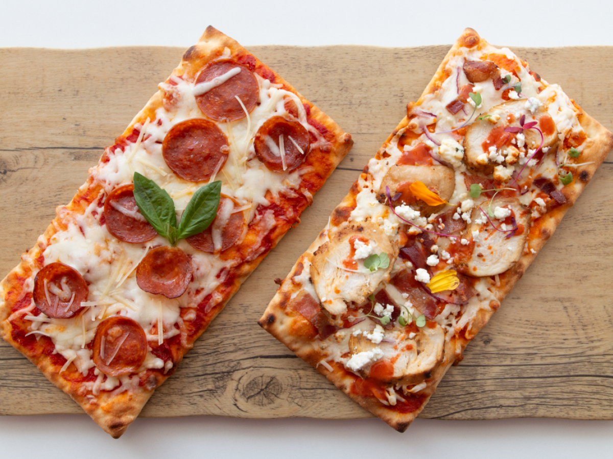 The image shows two rectangular pizzas on a wooden board, one with pepperoni and basil, the other with various toppings including chicken and vegetables.