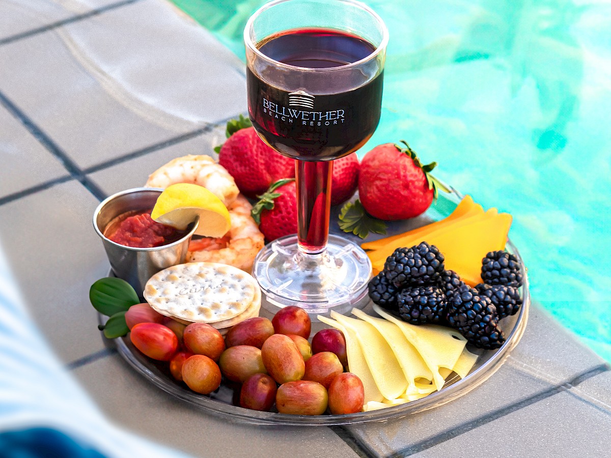 A platter with a glass of red wine, fruits, cheese, berries, and dips is placed by a poolside.