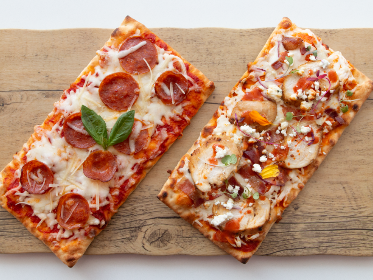 Two rectangular pizzas on a wooden board: one with pepperoni and basil; the other with chicken, bacon, red onions, and feta.