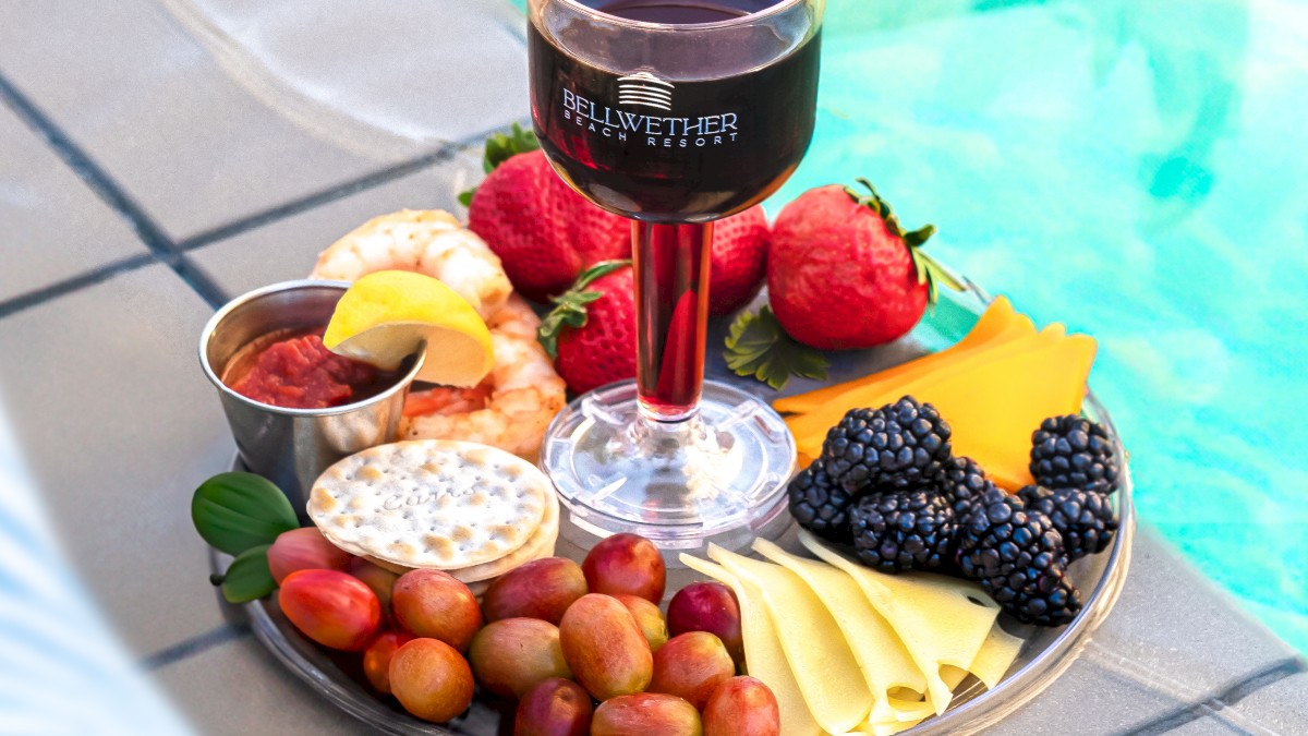 A platter with a glass of red wine, fruits, cheese, berries, and dips is placed by a poolside.