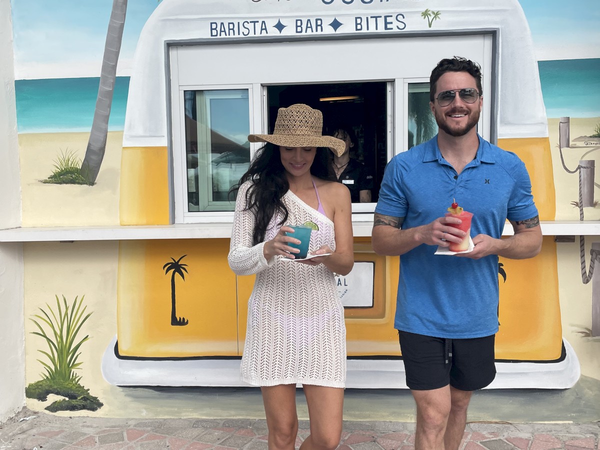 A woman and a man are standing in front of a beach-themed coffee kiosk, holding drinks. Palm trees and beach scene are painted on the kiosk backdrop.