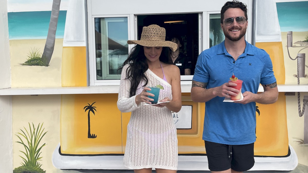 A woman and a man are standing in front of a beach-themed coffee kiosk, holding drinks. Palm trees and beach scene are painted on the kiosk backdrop.