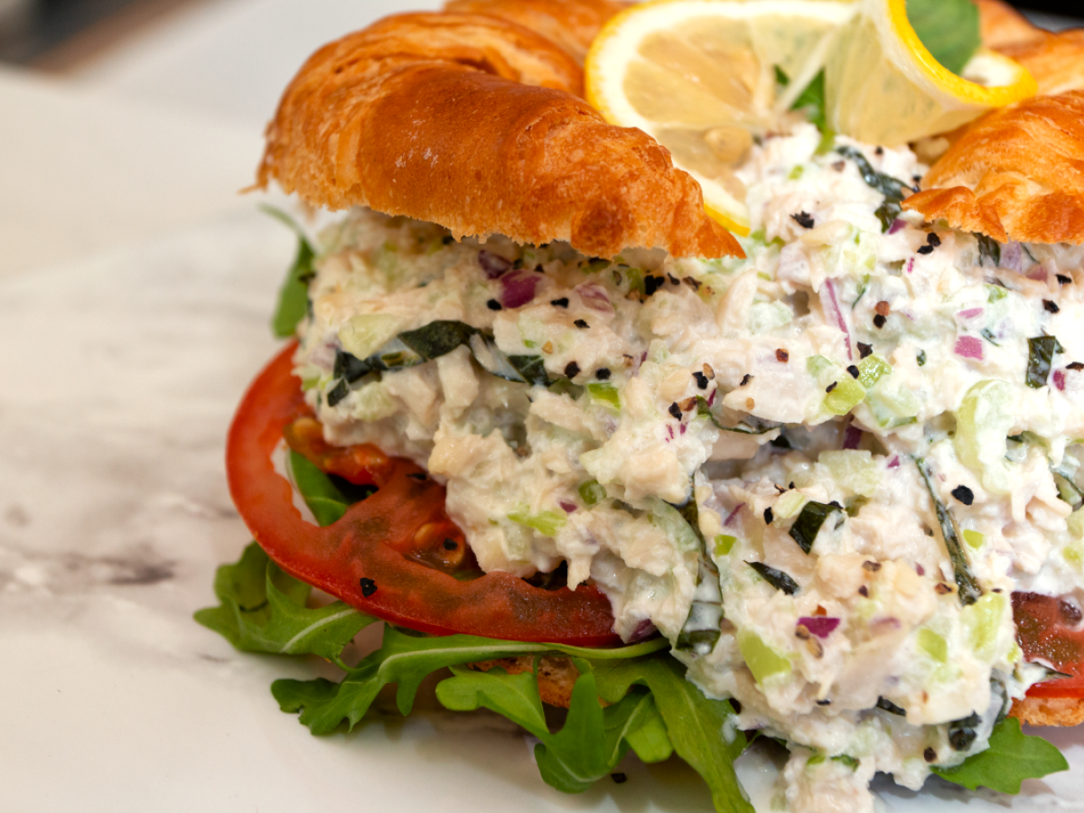 A croissant sandwich filled with a creamy salad mix, possibly chicken, with lettuce, tomato, and garnished with lemon slices on a marble surface.