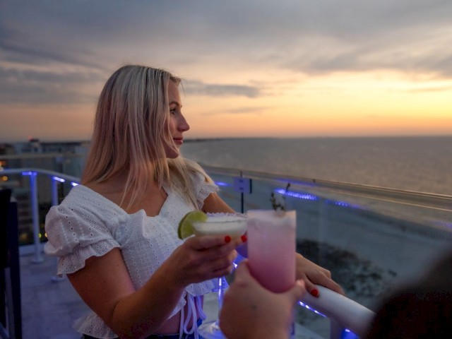 A woman with a white top holds a cocktail while enjoying a sunset view by the ocean, with another person also holding a drink.