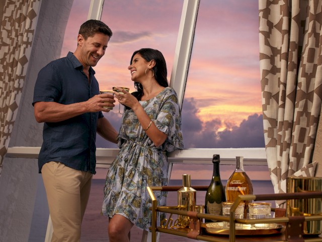 A couple is enjoying drinks together by the window during sunset. There are various bottles and glasses on a cart next to them.