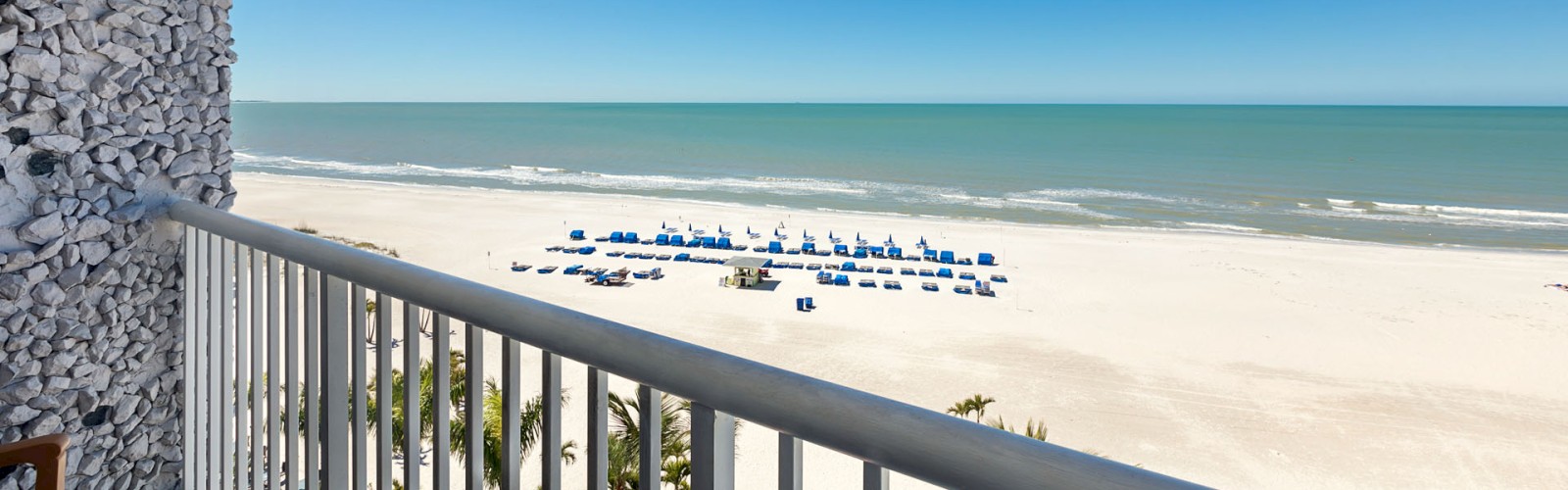 The image shows a view from a balcony overlooking a sandy beach with rows of blue umbrellas, calm ocean waves, and a clear blue sky.