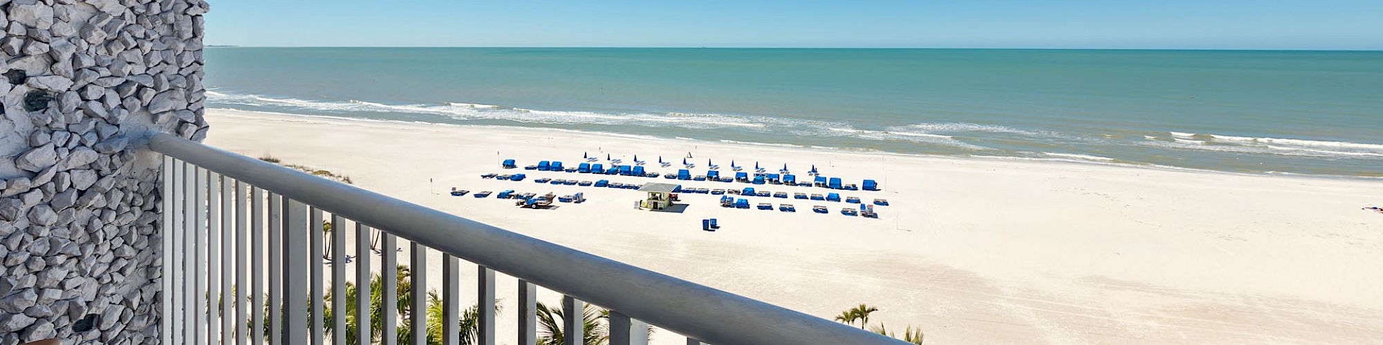 The image shows a view from a balcony overlooking a sandy beach with rows of blue umbrellas, calm ocean waves, and a clear blue sky.