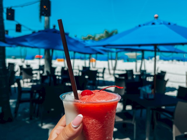 A hand holds a layered frozen drink with a straw. The background shows a beach setting with tables, chairs, and blue umbrellas.